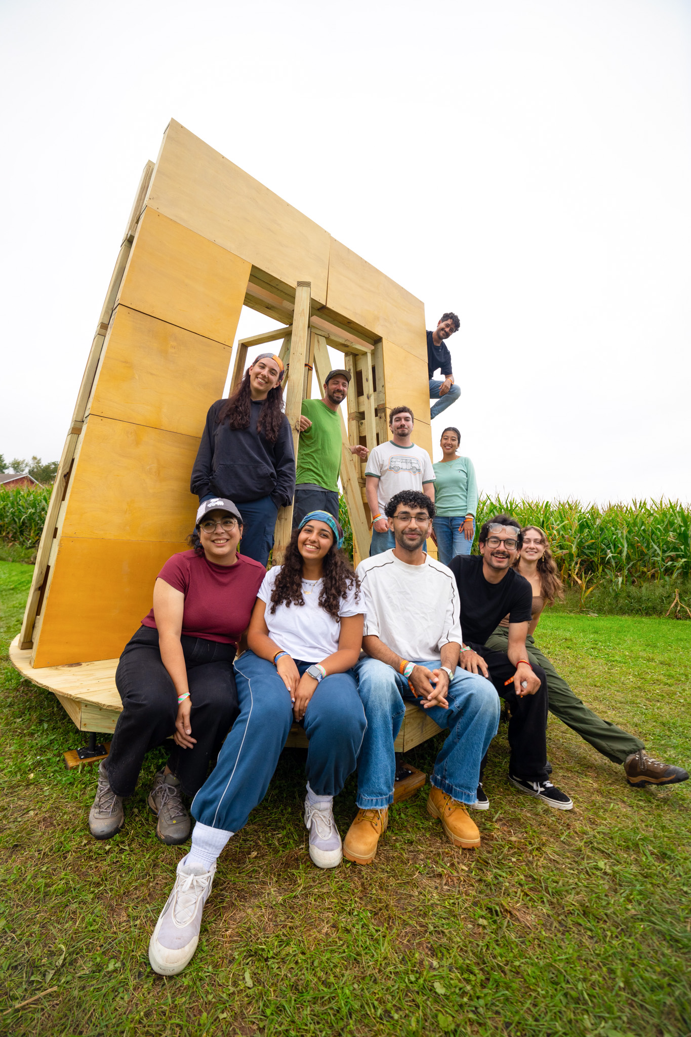 The faculty leads and student build team seated on the platform in front of the plywood frame, with a cornfield behind